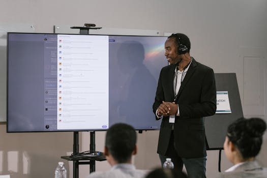 An adult man presents at a technology seminar using a large screen and headset.